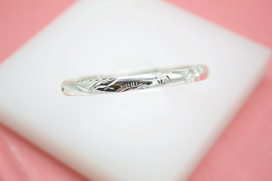 A silver bangle bracelet with intricate engraved patterns rests horizontally on a white display block against a soft pink background.