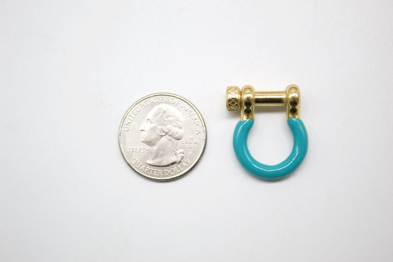 A shiny gold and turquoise U-shaped screw shackle rests next to a US quarter coin to show scale on a white surface.