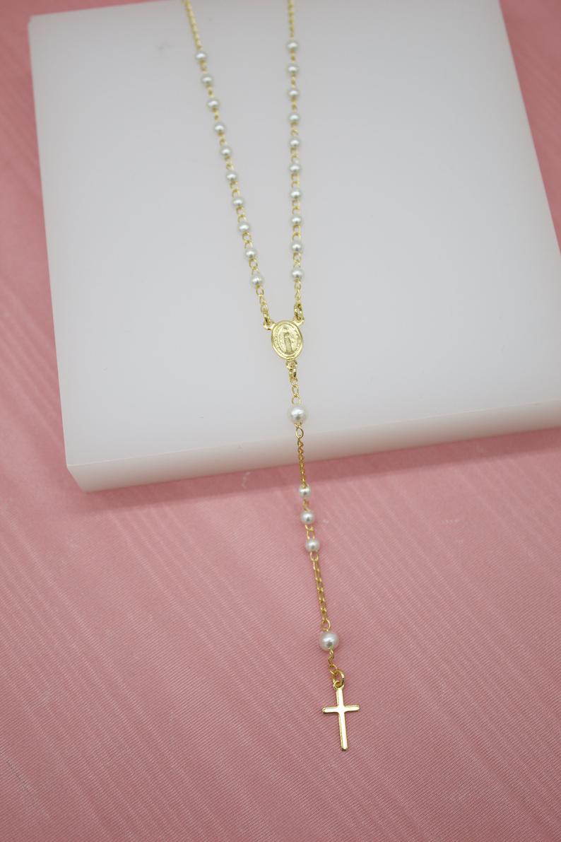 A delicate gold rosary necklace featuring white pearl beads and a hanging cross pendant rests on a white display block.