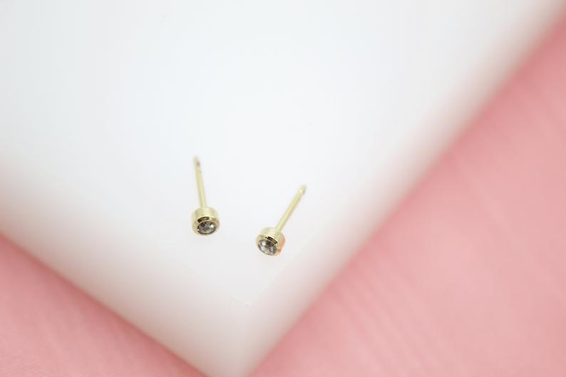 A pair of delicate gold bezel-set stud earrings rests on a white surface against a pink background.