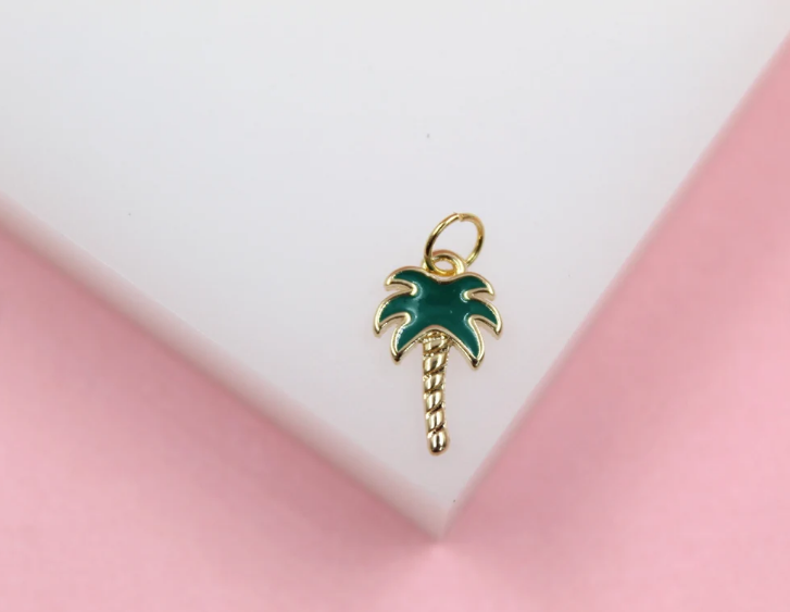 A small gold palm tree charm featuring vibrant green enamel leaves rests on a white display block against a soft pink background.