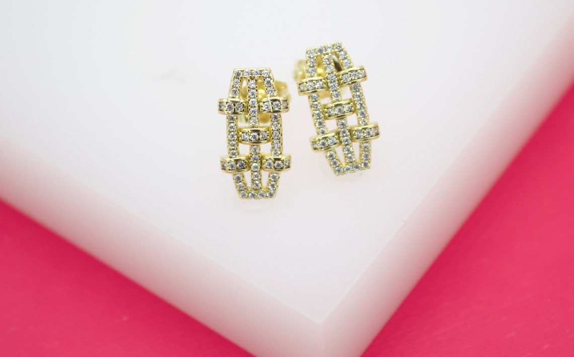 A pair of geometric gold earrings encrusted with sparkling pave diamonds rests on a white and pink display surface.