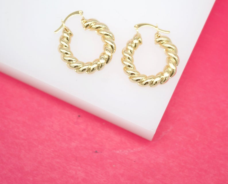 A pair of glossy gold twisted hoop earrings rests on a white platform against a pink background.