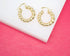 A pair of glossy gold twisted hoop earrings rests on a white platform against a pink background.