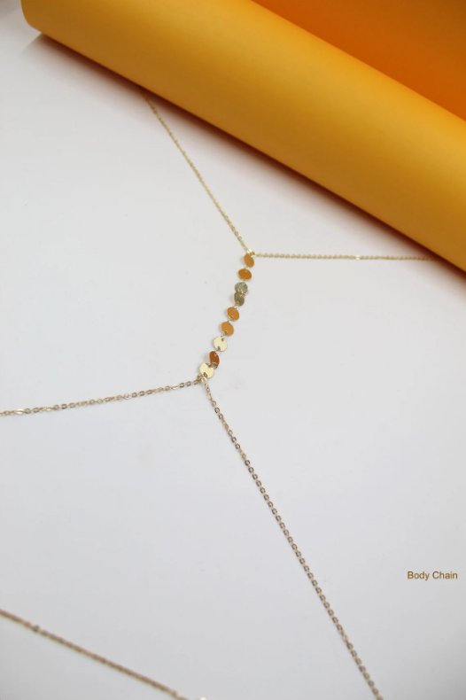 A delicate gold body chain rests on a white surface beneath a bright orange paper roll.