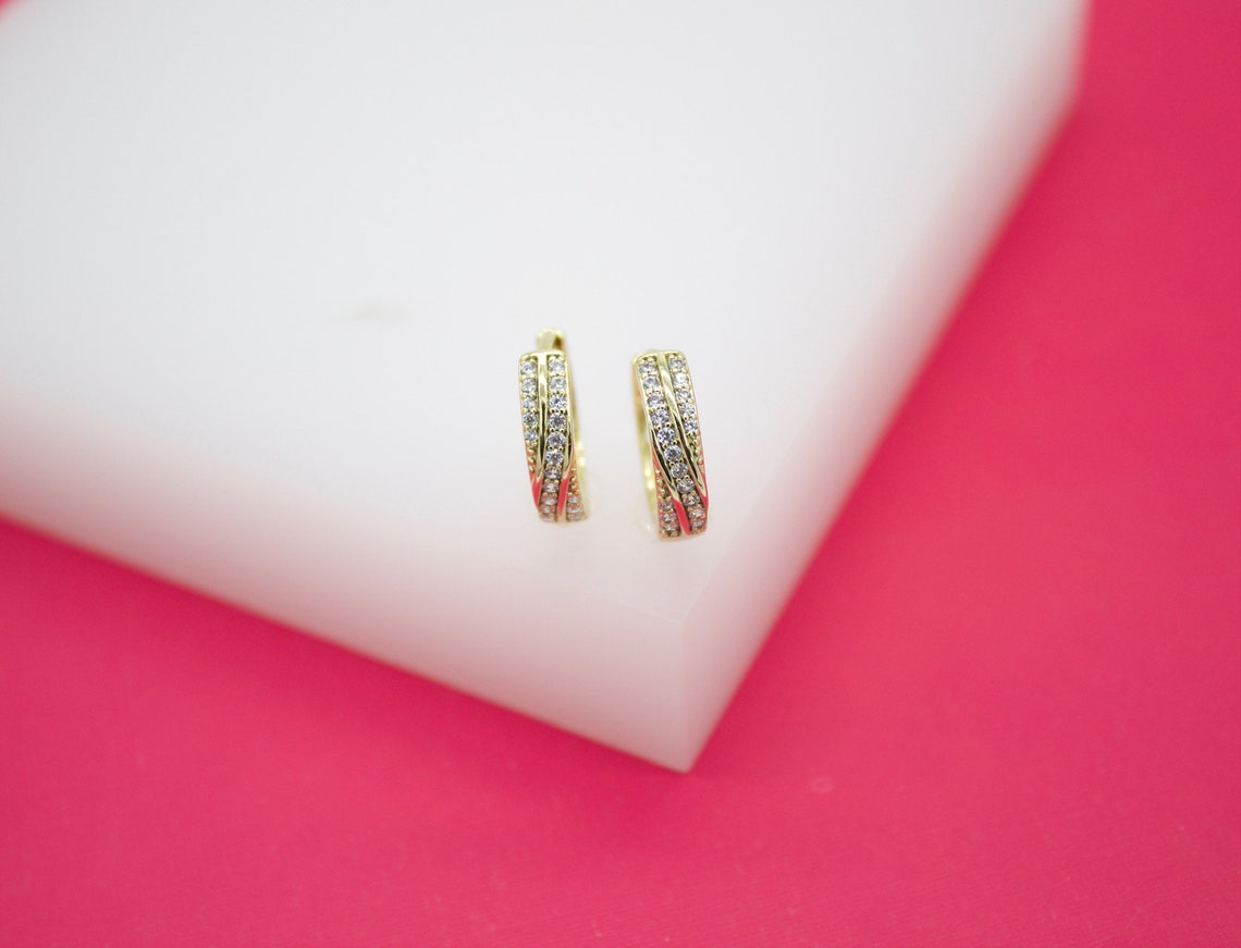 A pair of gold huggie earrings featuring sparkling pave-set cubic zirconia stones rests on a white display block.