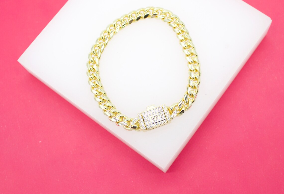 A gold cuban link bracelet rests on a white display stand against a vibrant pink background.