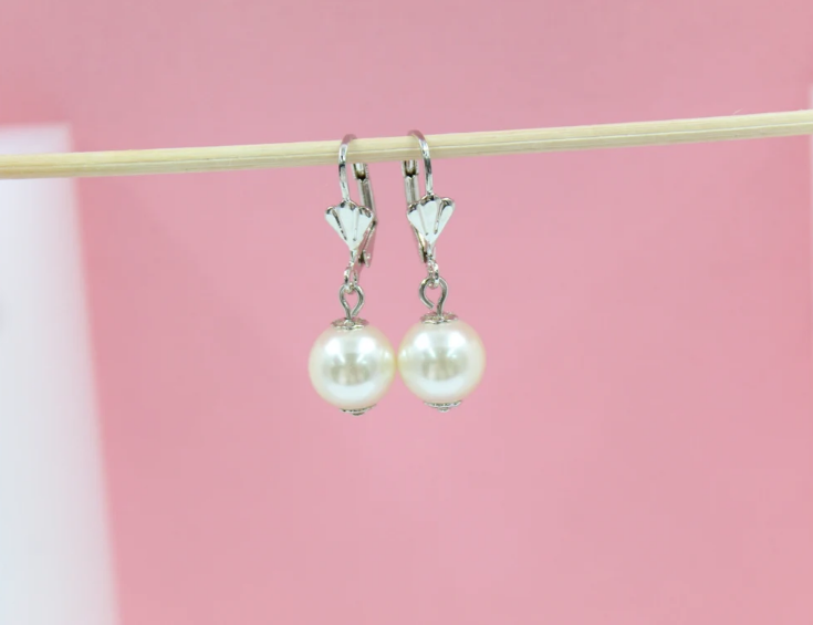 A pair of elegant silver leverback pearl earrings hang from a wooden stick against a soft pink background.