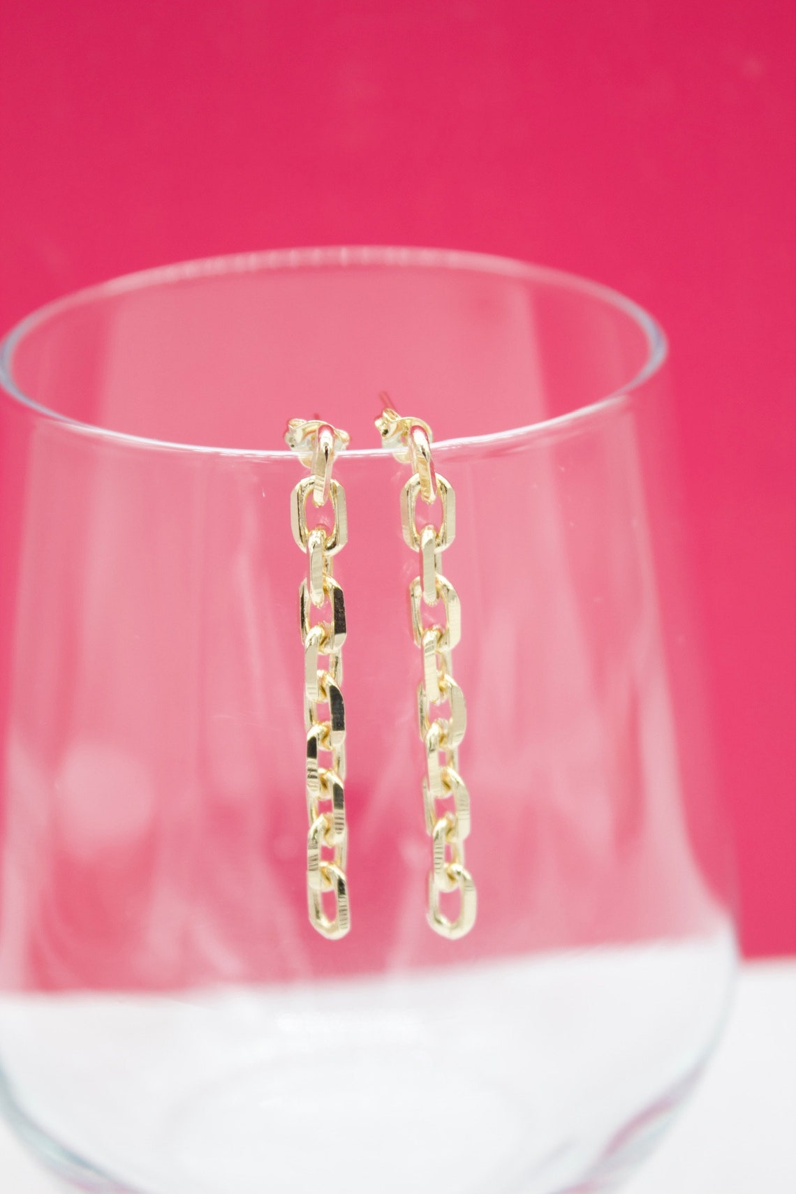 A pair of dangling gold chain-link earrings rests on the rim of a clear glass against a vibrant pink background.