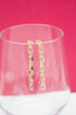 A pair of dangling gold chain-link earrings rests on the rim of a clear glass against a vibrant pink background.
