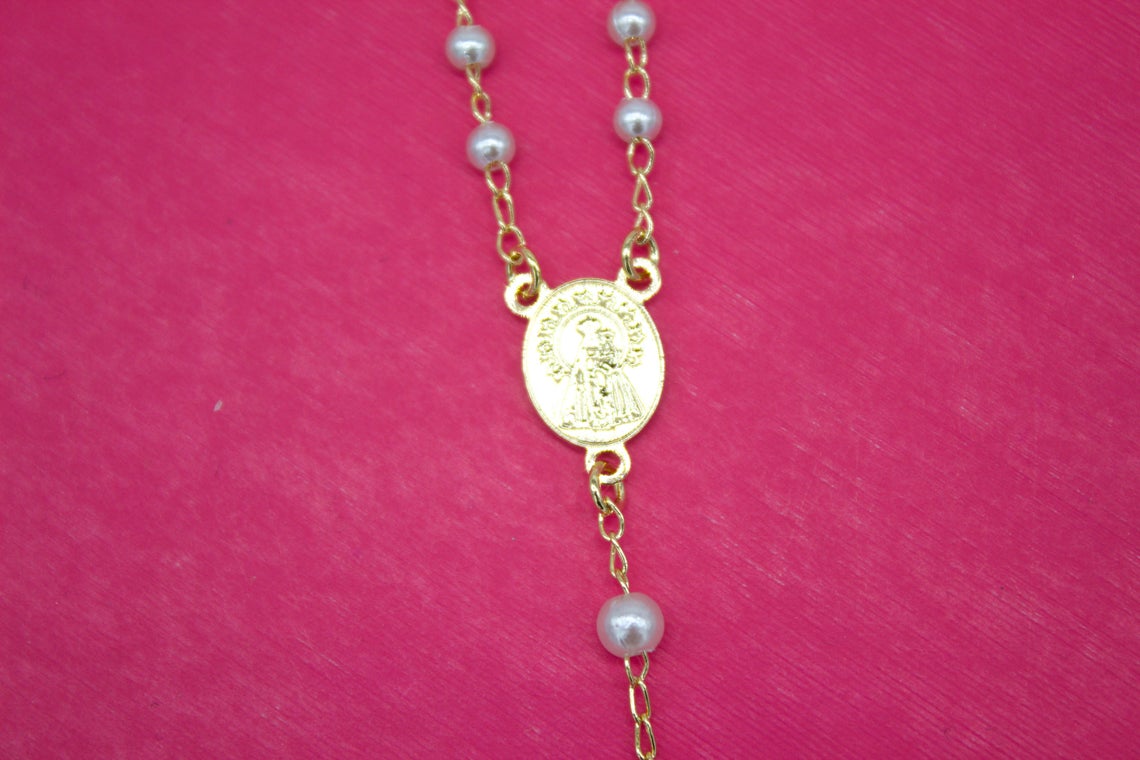 A delicate gold rosary necklace with white pearl beads and a central religious medallion rests against a vibrant pink background.