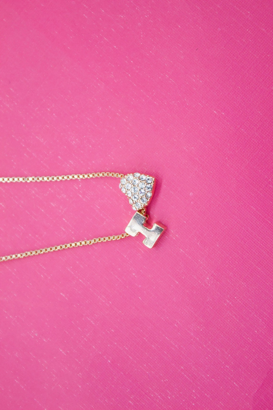 A delicate gold chain necklace featuring a rhinestone-encrusted heart and initial pendant rests against a textured hot pink background.
