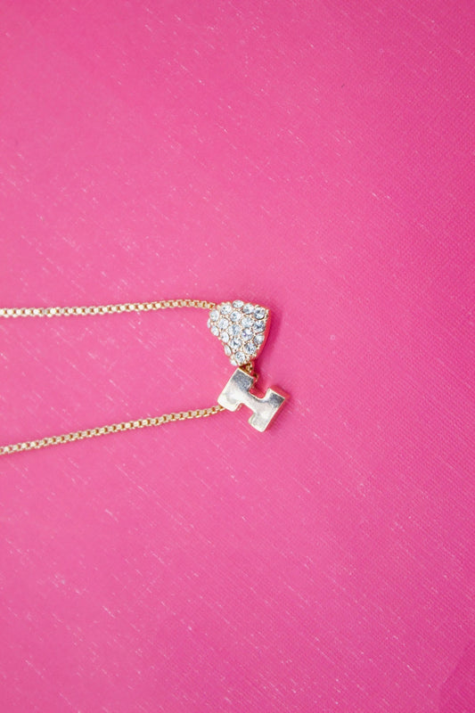 A delicate gold chain necklace featuring a rhinestone-encrusted heart and initial pendant rests against a textured hot pink background.