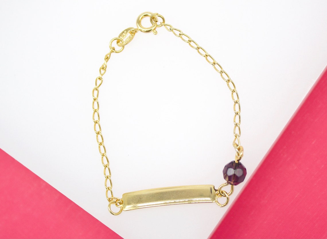 A delicate gold ID bracelet rests against a white and pink background, featuring a polished engravable identification plate alongside a dangling deep purple faceted bead.