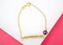 A delicate gold ID bracelet rests against a white and pink background, featuring a polished engravable identification plate alongside a dangling deep purple faceted bead.