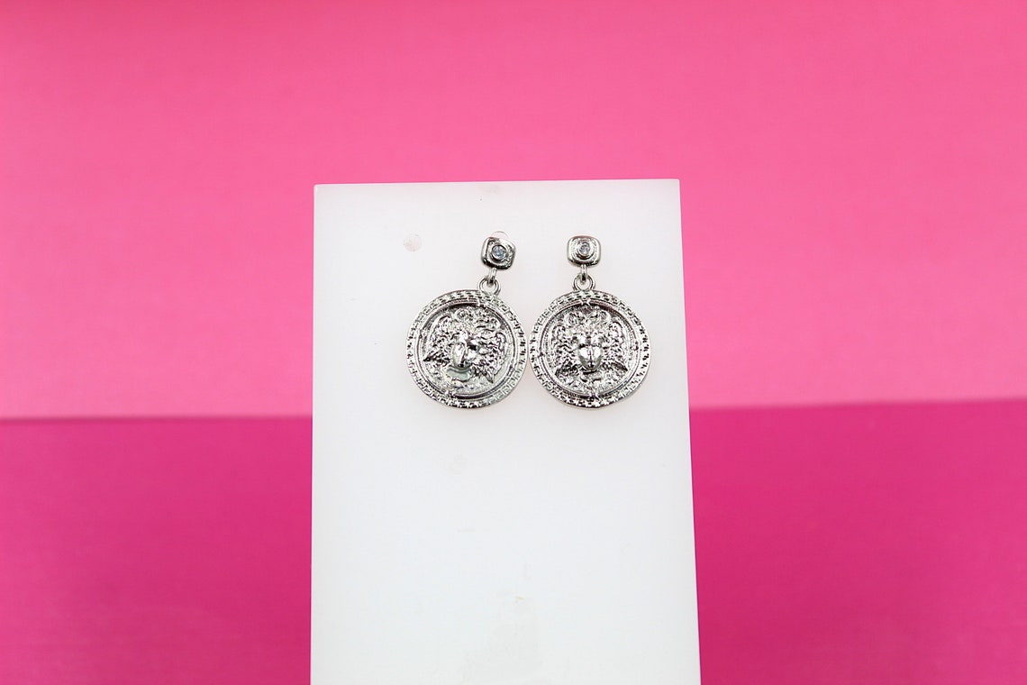 A pair of intricate silver coin earrings featuring a detailed lion head design hangs on a white display stand against a pink background.