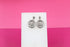 A pair of intricate silver coin earrings featuring a detailed lion head design hangs on a white display stand against a pink background.