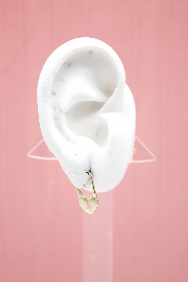 A white ear model displays a gold heart-shaped padlock earring against a solid pink background.