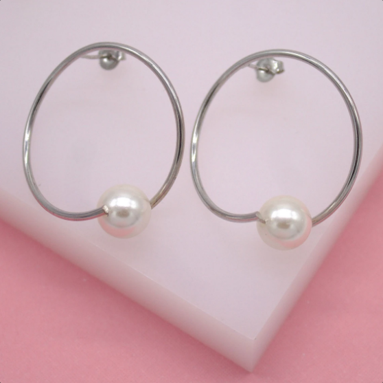 A pair of modern silver hoop earrings featuring a single dangling white pearl displayed on a pink and white surface.