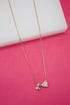 A delicate gold chain necklace drapes against a vibrant pink background, featuring a charm with the letter H alongside a sparkling rhinestone-embossed heart pendant.