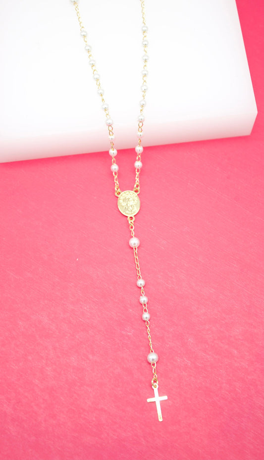 A delicate gold rosary necklace featuring white faux pearl beads and a small crucifix rests against a vibrant pink and white background.