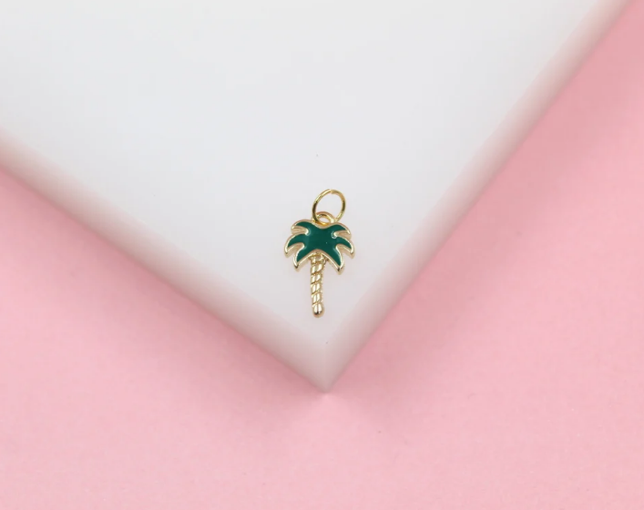 A small gold palm tree pendant featuring green enamel leaves rests on a white and pink surface.
