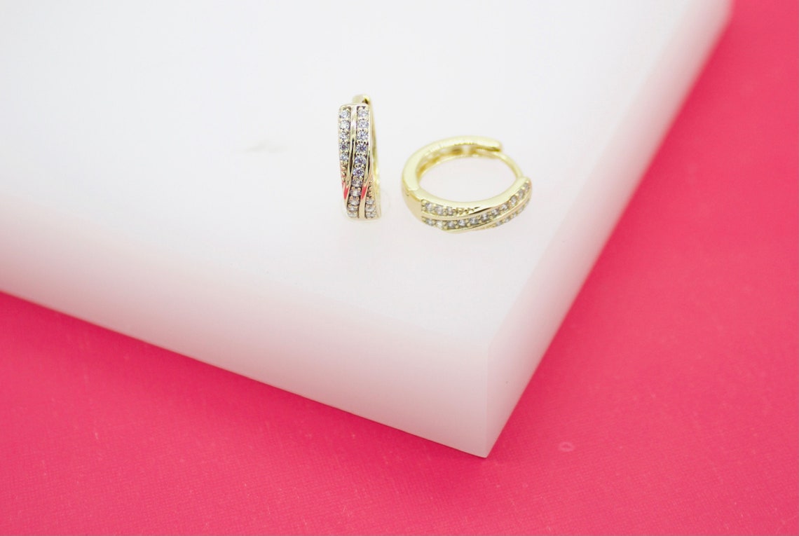 A pair of gold huggie hoop earrings embedded with sparkling cubic zirconia stones rests on a white display surface against a vibrant pink background.