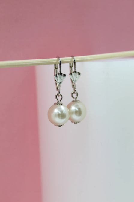 A pair of elegant faux pearl drop earrings with silver leverback closures hangs from a thin wooden stick against a pink and white backdrop.