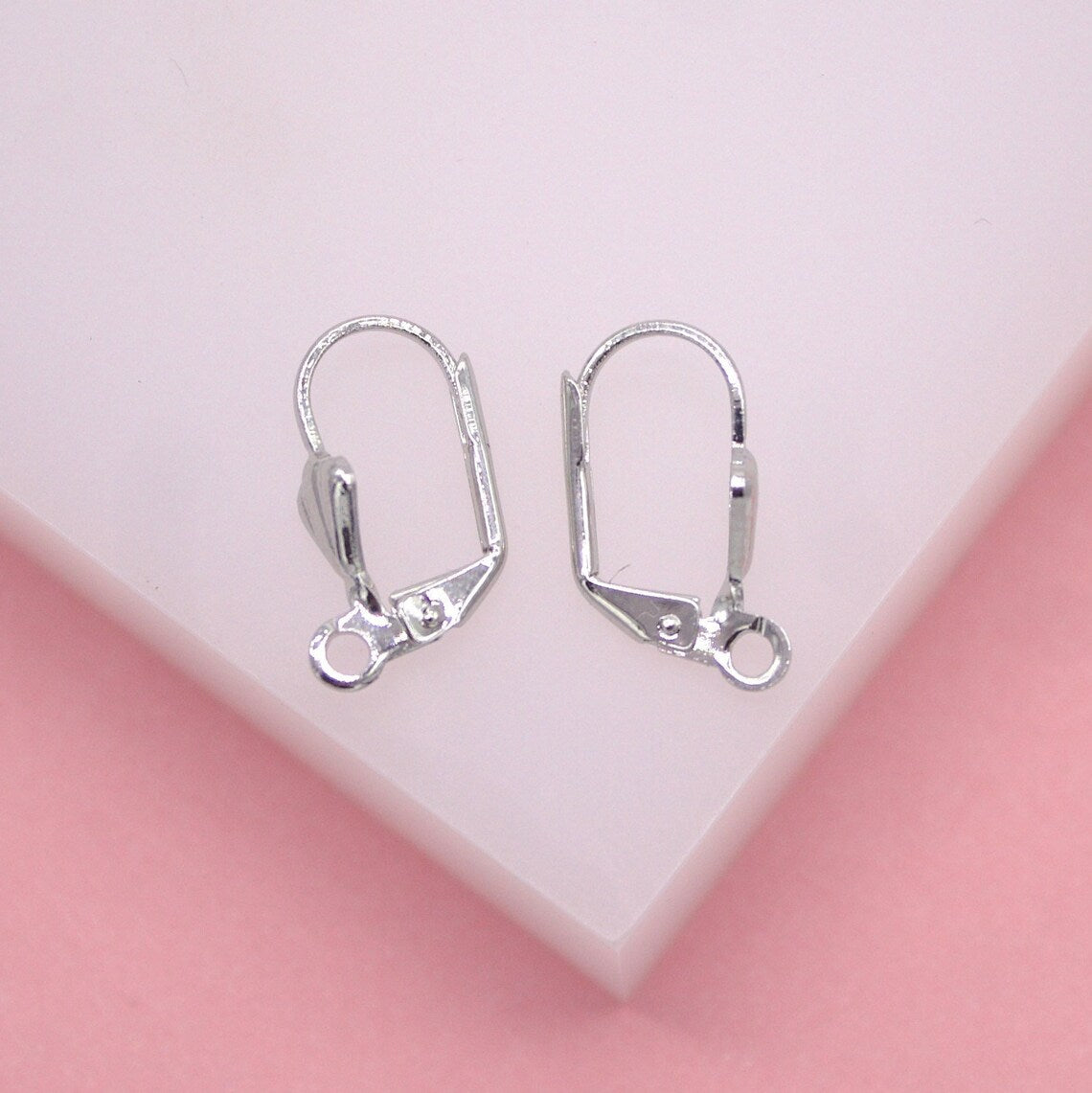 A pair of shiny silver leverback earring findings rests on a soft pink surface.