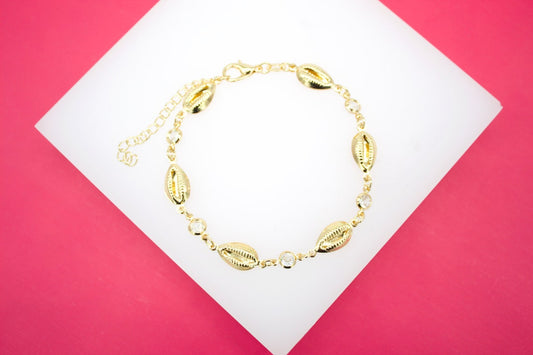 A delicate gold seashell bracelet featuring sparkling cubic zirconia stones rests on a white display against a vibrant pink background.