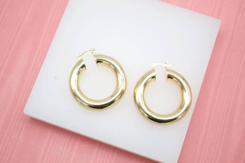 A pair of polished gold hoop earrings rests neatly on a white display surface against a pink background.