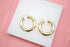 A pair of polished gold hoop earrings rests neatly on a white display surface against a pink background.