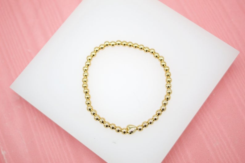 A shiny gold bead stretch bracelet rests on a white display against a pink background.