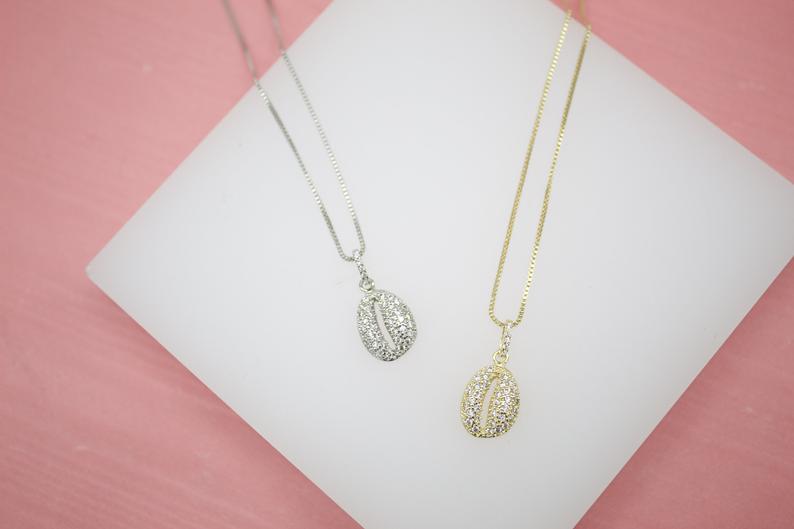 A silver and a gold coffee bean pendant necklace sparkle on a white display block against a soft pink background.
