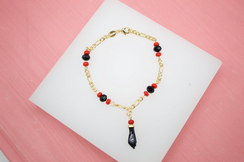 A gold chain bracelet features alternating red and black crystal beads and a black figa charm charm resting on a white display block.