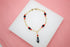 A gold chain bracelet features alternating red and black crystal beads and a black figa charm charm resting on a white display block.