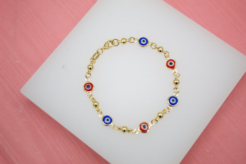 A delicate gold evil eye bracelet featuring alternating blue and red enamel evil eye charms rests on a white display surface.