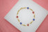 A delicate gold evil eye bracelet featuring alternating blue and red enamel evil eye charms rests on a white display surface.