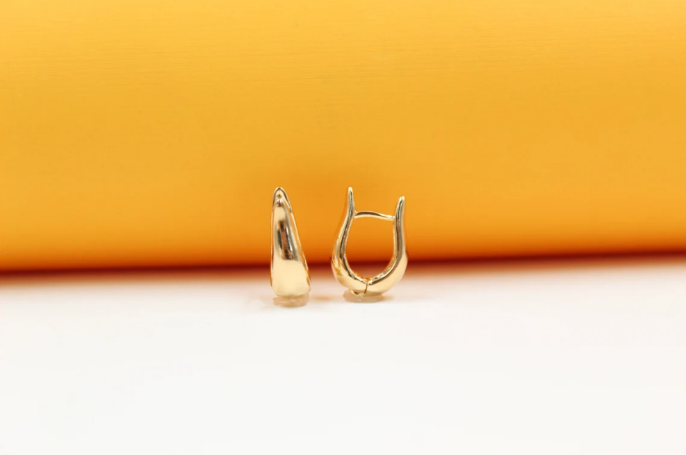A pair of sleek gold huggie earrings rests against a vibrant orange and white background.