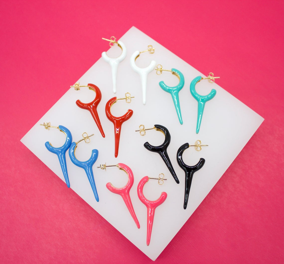 A colorful collection of enamel and gold hoop earrings with pointed charms is arranged neatly on a white display surface against a vibrant pink background.