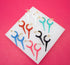 A colorful collection of enamel and gold hoop earrings with pointed charms is arranged neatly on a white display surface against a vibrant pink background.