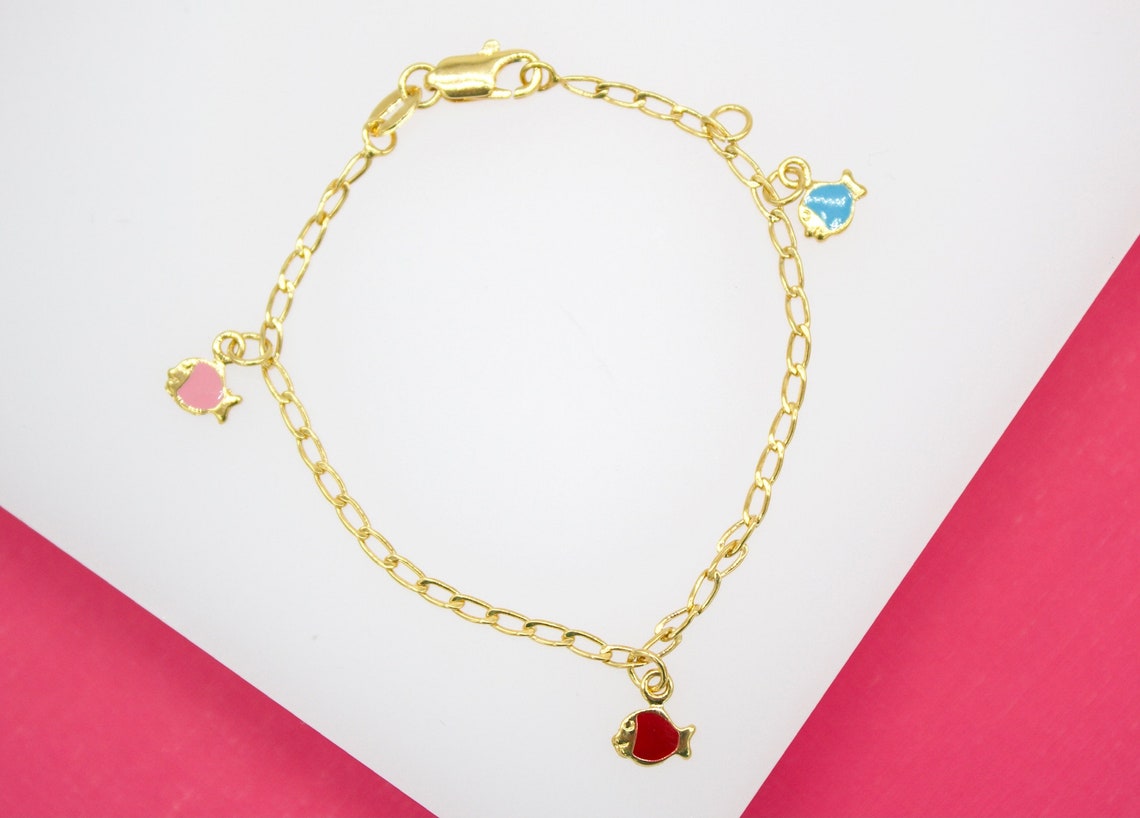A delicate gold-plated children's charm bracelet displays three colorful enamel fish charms in pink, blue, and red against a white and pink background.