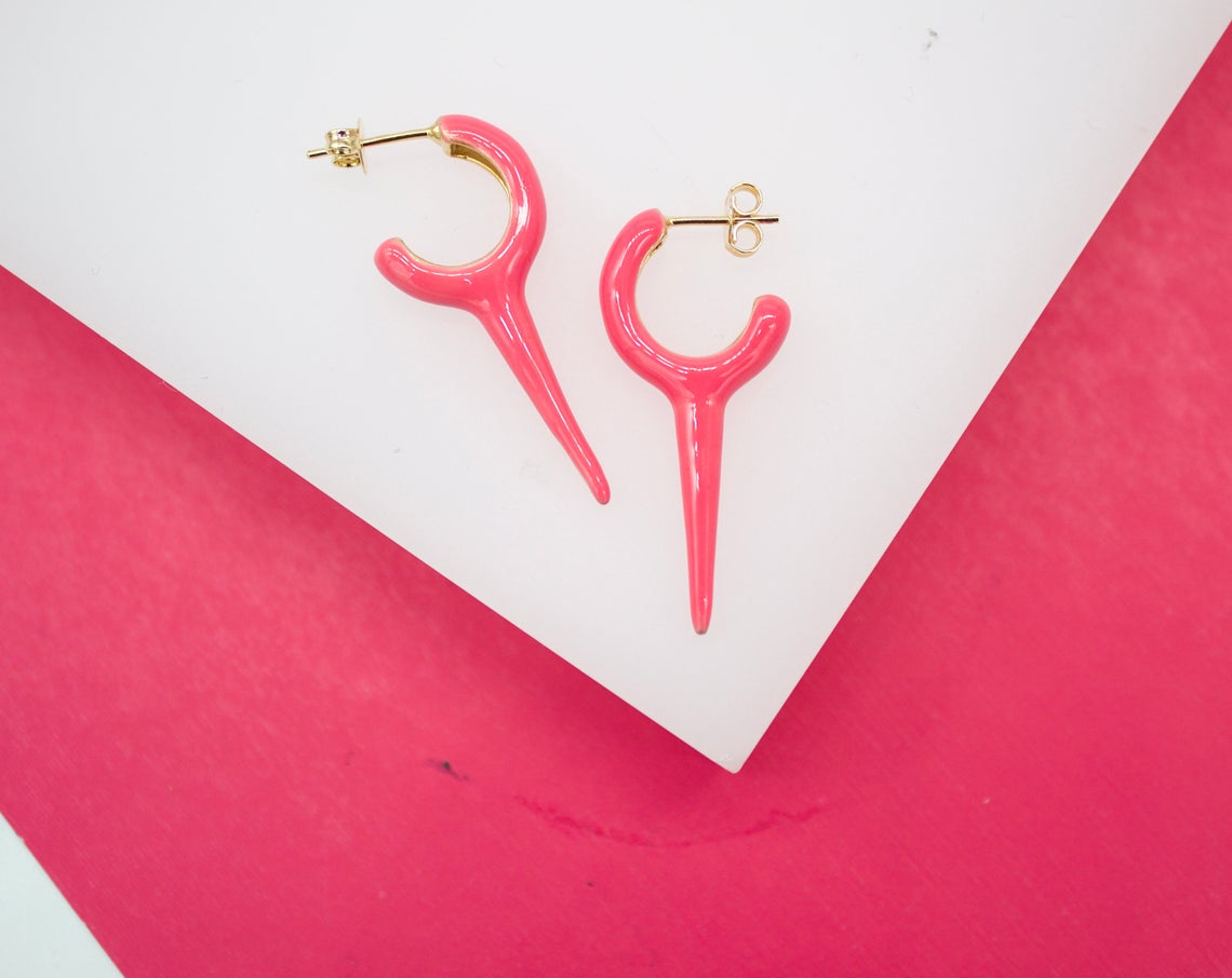 A pair of vibrant pink enamel hoop earrings with dangling dagger charms rests on a white and bright pink background.