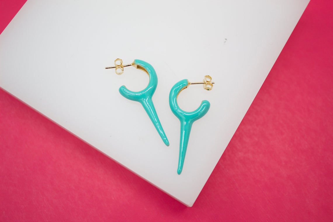 A pair of vibrant turquoise enameled huggie hoop earrings with pointed dagger drops rests on a white display against a bright pink background.