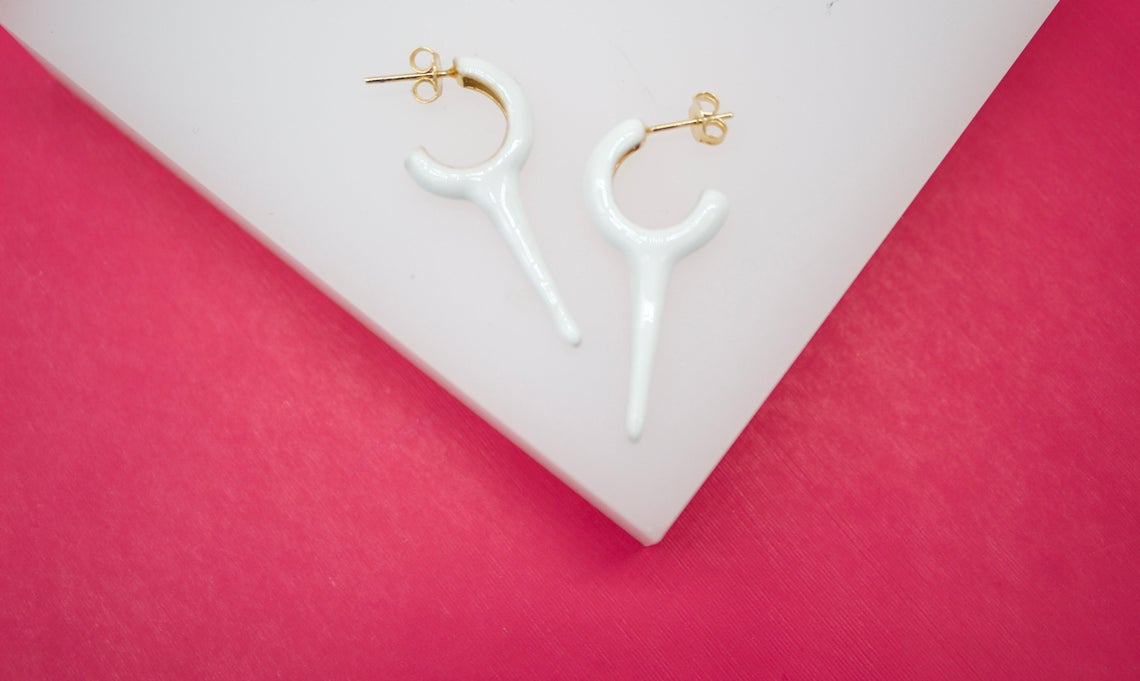 Two glossy white hoop earrings with dangling spike charms rest on a white display against a vibrant pink background.