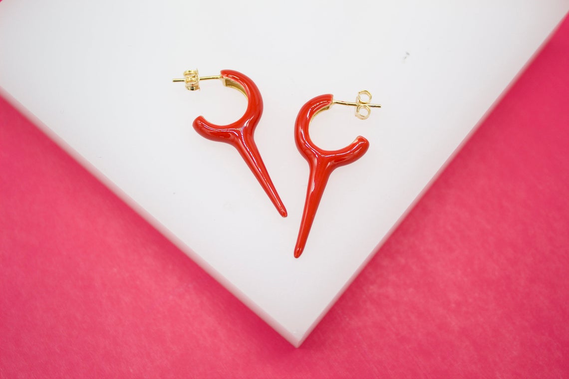 Two glossy red horn-shaped hoop earrings rest on a white surface against a vibrant pink background.