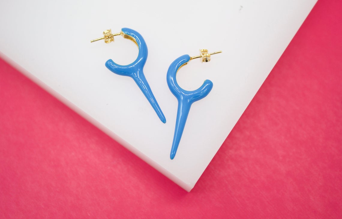 A pair of vibrant blue enamel hoop earrings with a dangling spike design rests on a white surface against a bright pink background.