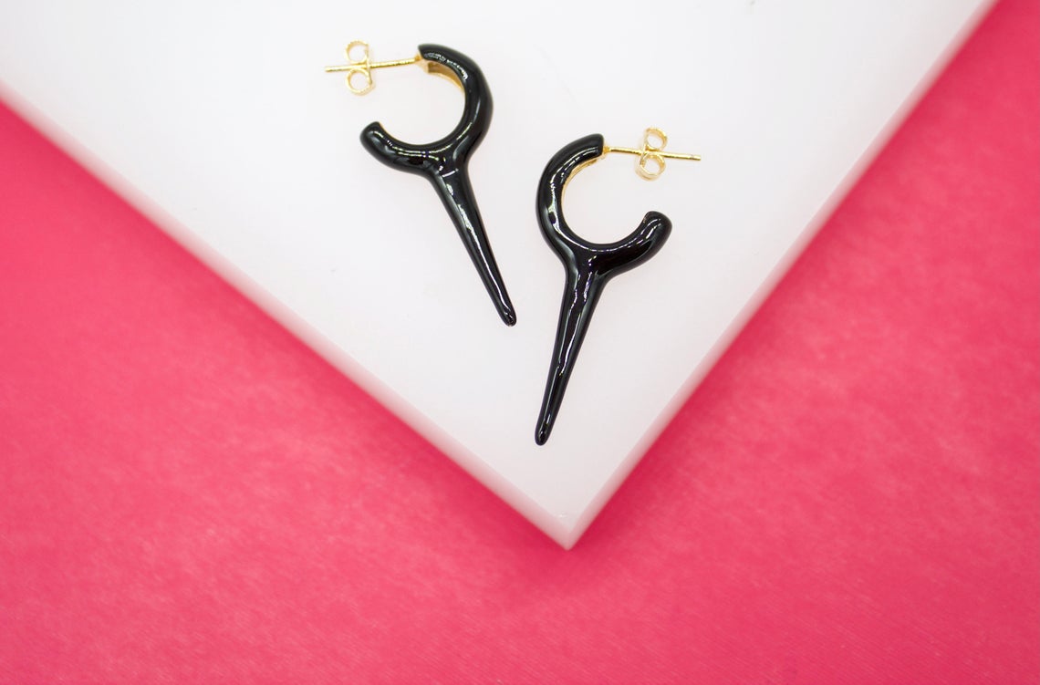 A pair of glossy black spiked hoop earrings rests on a white platform against a pink background.