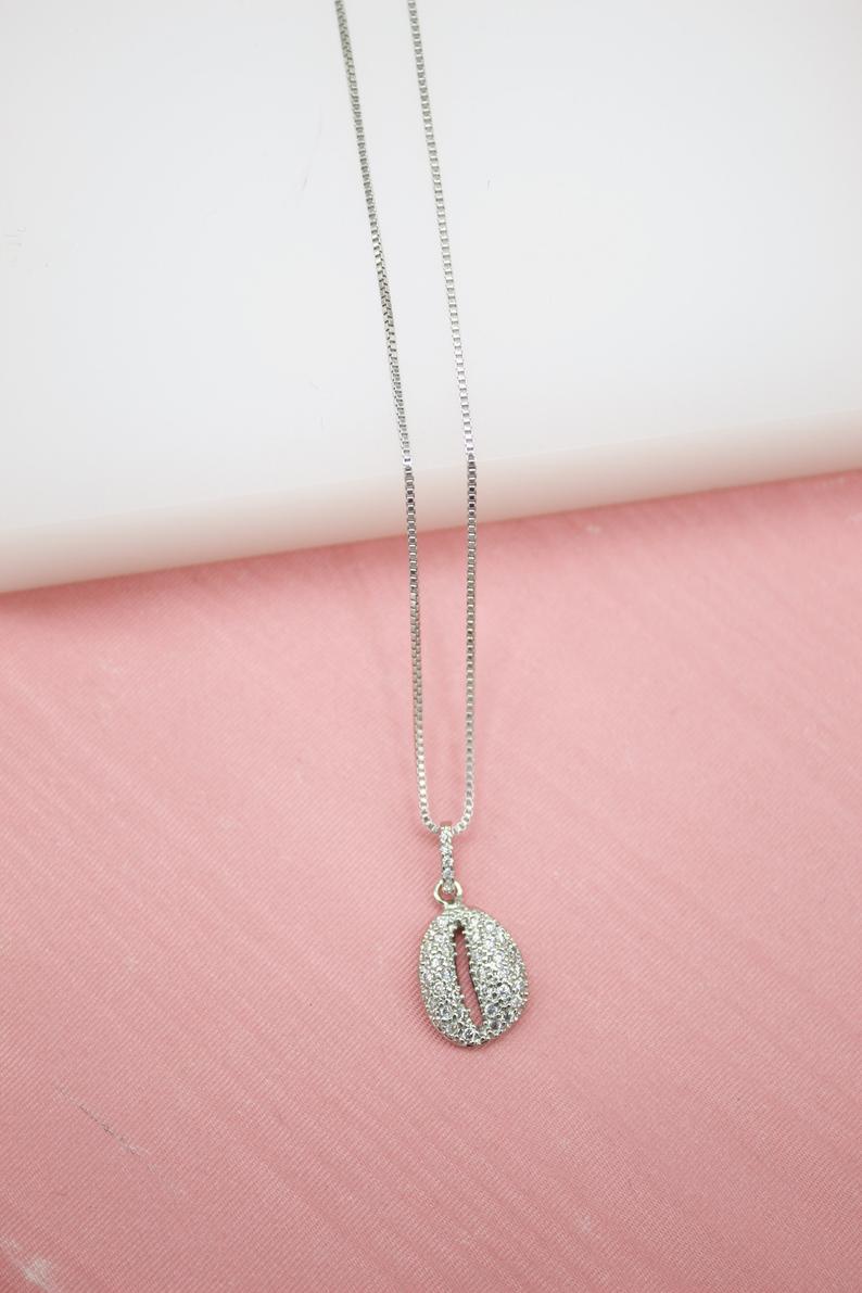 A delicate silver box chain necklace features a sparkling pavé-set cowrie shell pendant displayed against a soft pink and white background.