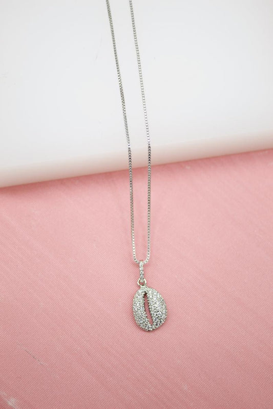 A delicate silver box chain necklace features a sparkling pavé-set cowrie shell pendant displayed against a soft pink and white background.